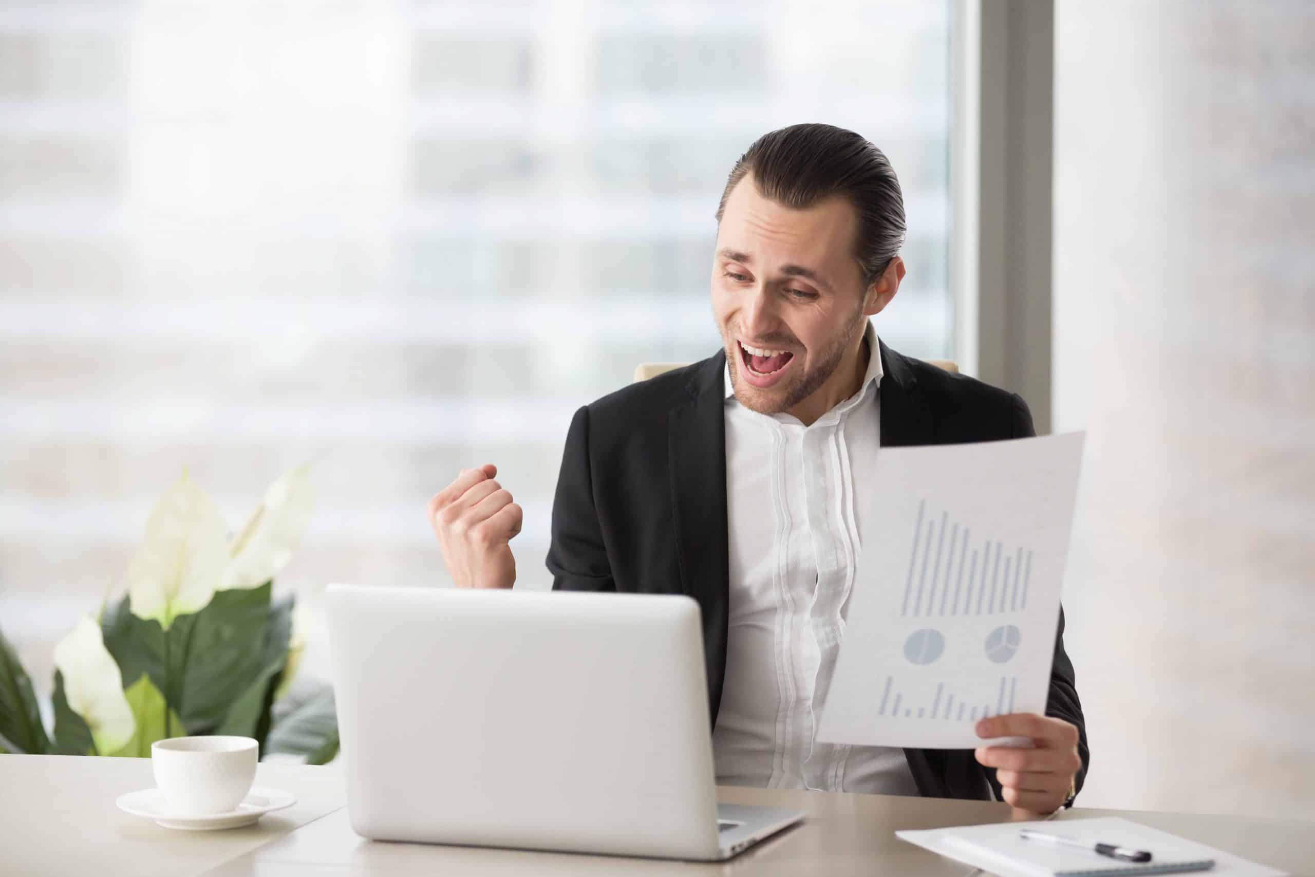 Happy handsome project manager in office celebrates and screams with happiness, looking at laptop screen and holding positive financial report. Great news on stock investment, finance victory concept.